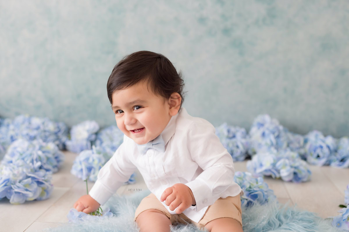 Witness the charm of a 11-month-old boy, donning a white shirt paired with a blue bow, seated on a cozy blue rug against a serene blue backdrop adorned with delicate blue flowers. This captivating moment was elegantly captured during a toddler photoshoot in Delhi, Gurgaon, and Noida by the skilled photographer, Anega Bawa.