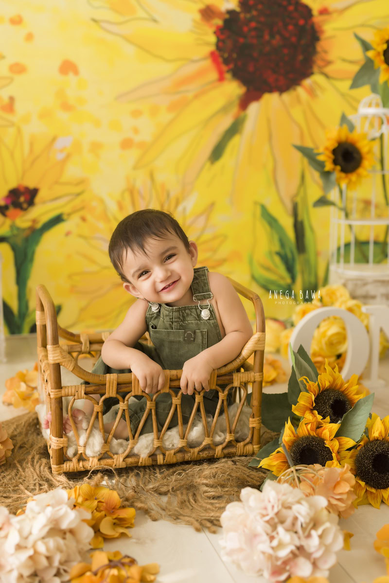 Envision the enchanting sight of an 11-month-old boy nestled in a wooden cot, surrounded by warmth from a brown rug, all against a backdrop adorned with vibrant sunflowers. This captivating moment was elegantly captured during a toddler photoshoot in Delhi, Gurgaon, and Noida by the skilled photographer, Anega Bawa.