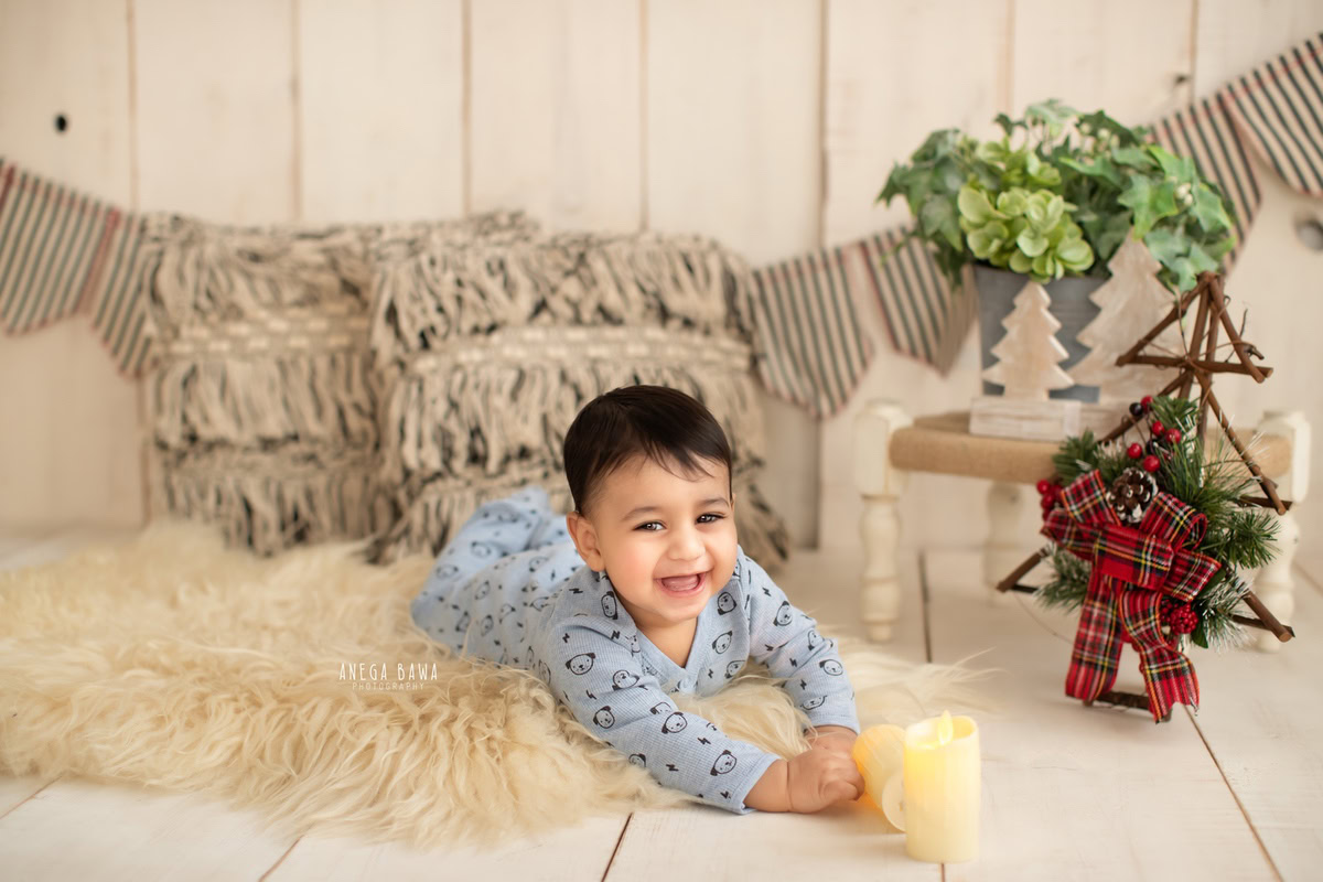During this charming sitter photo shoot in Delhi, Gurgaon by Anega Bawa, a 12-month-old boy lies comfortably on a beige rug surrounded by cushions. Against a soothing beige backdrop, candles add warmth and ambiance to the scene, creating a serene atmosphere for capturing this precious moment.
