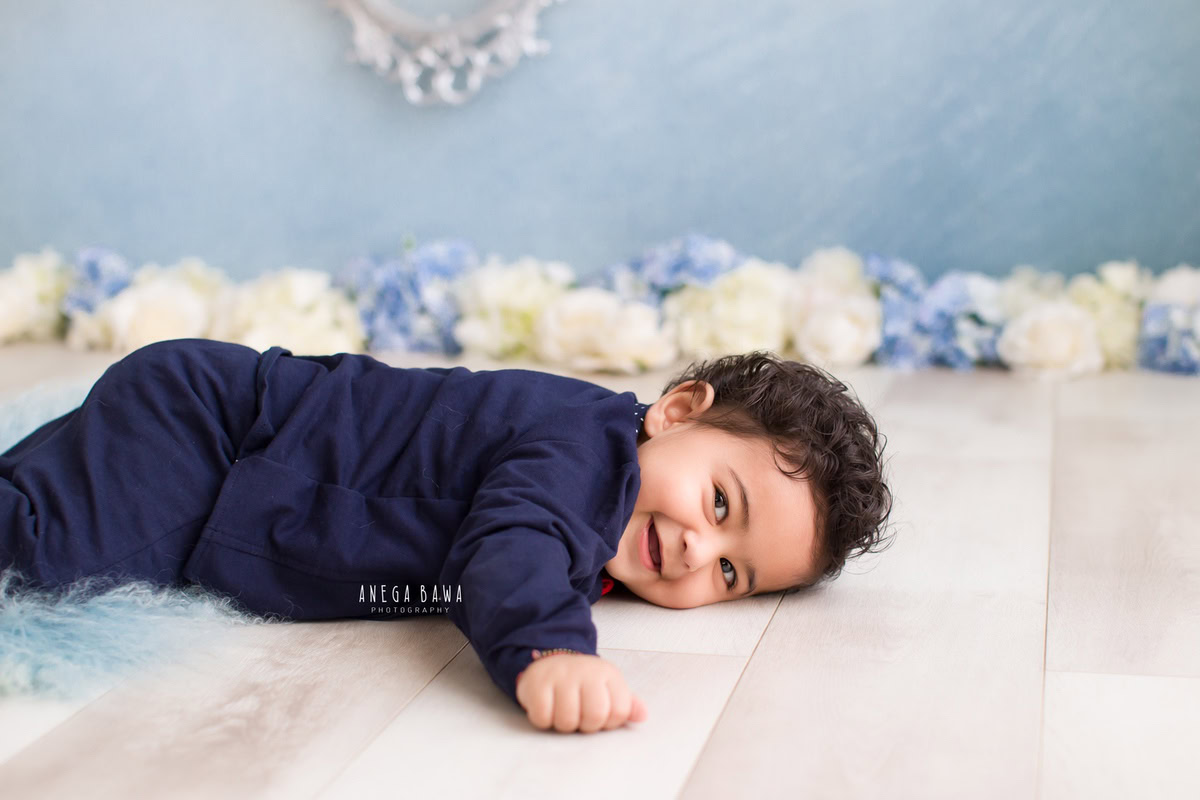 Capture the precious moments of your little boy's growth with Anega Bawa Photography's sitter photoshoot in Delhi and Gurgaon. Dressed in a cute blue babysuit, he lies against a serene blue backdrop adorned with blue and white flowers, marking his 12-month milestone beautifully.