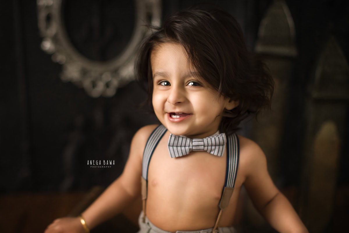 In this delightful sitter photo session in Delhi, Gurgaon by Anega Bawa, the mischievous spirit of a 12-month-old boy shines through as he strikes a playful pose against a rich brown backdrop. With a cute bow adorning his outfit, he adds an extra touch of charm to the frame.