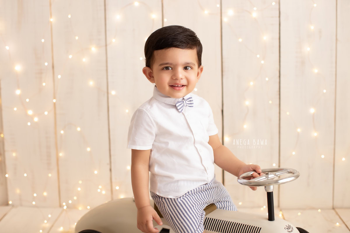 Celebrate your little boy's 12-month milestone with a charming sitter photoshoot by Anega Bawa Photography in Delhi and Gurgaon. Watch him joyfully ride a toy scooter against a warm beige backdrop, illuminated by golden lights, capturing his playful spirit in every frame.
