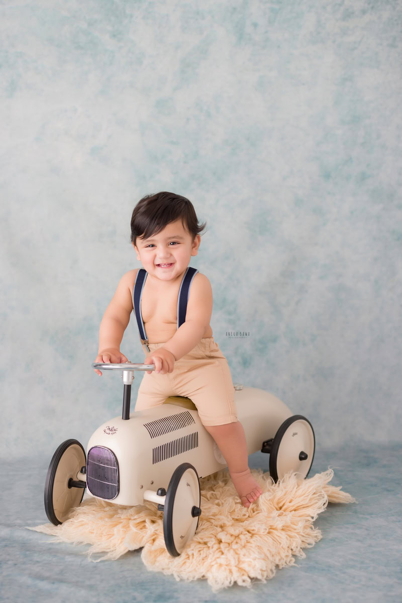 Let's envision your little explorer at Anega Bawa Photography's sitter photoshoot in Delhi and Gurgaon. Imagine him joyfully riding a toy scooter on a beige rug, with a vibrant blue backdrop adding a pop of color to the scene. These delightful moments freeze in time the spirit of adventure and discovery at 12 months old.