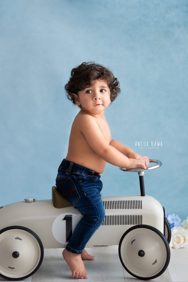 Captured by sitter photographer Anega Bawa in Delhi, a joyful 12-month-old boy gleefully rides a toy scooter against a vibrant blue backdrop.