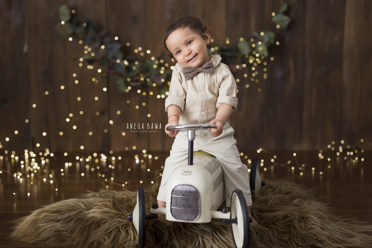 Charming boy riding a toy scooter on a cozy brown rug surrounded by twinkling fairy lights against a warm brown backdrop, celebrating his 12 months first birthday photoshoot in Delhi by Anega Bawa, Gurgaon, Noida
