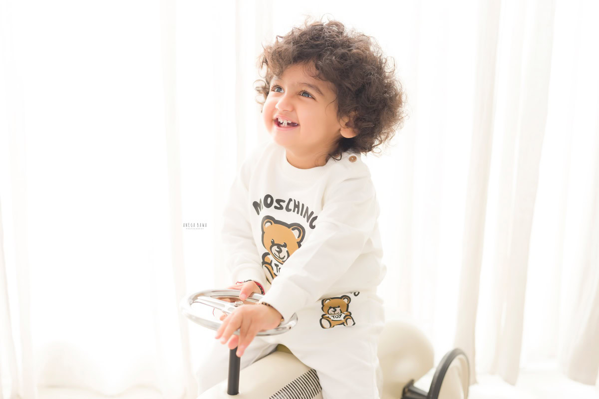 Captured during a delightful baby pre-birthday photoshoot by Anega Bawa in Delhi, this charming image features a 12-month-old boy with curly hair joyfully riding a toy scooter. Set against a clean white backdrop, the scene exudes a sense of innocence and playful energy, making it a memorable moment to cherish for years to come.