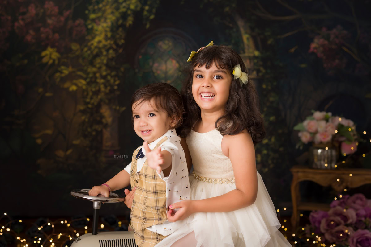 Adorable boy joyfully riding a toy scooter alongside his sibling, set against a warm brown backdrop illuminated by fairy lights, commemorating his 12 months first birthday photoshoot in Delhi by Anega Bawa, Gurgaon, Noida