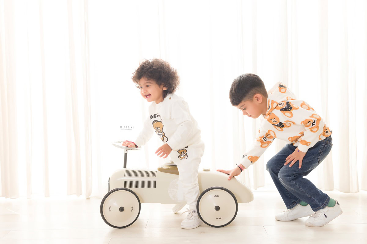 In this captivating image from a baby pre-birthday photoshoot by Anega Bawa in Delhi, a 12-month-old boy gleefully rides a toy scooter while accompanied by a sibling. Against a pristine white backdrop, their playful interaction is beautifully captured, radiating warmth and joy. It's a precious moment frozen in time, perfect for treasuring memories of their early years.