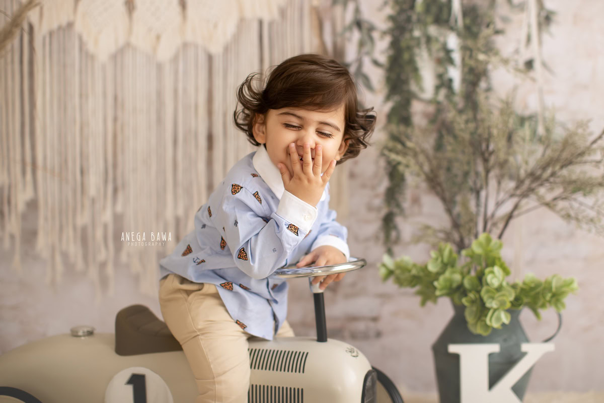 Capture the precious moments of your 12-month-old boy's journey with a delightful sitter photoshoot by Anega Bawa Photography in Delhi and Gurgaon. Let him zoom around on a toy scooter against a clean white backdrop, accentuated by leafy green pots, creating a whimsical and vibrant atmosphere for unforgettable memories.