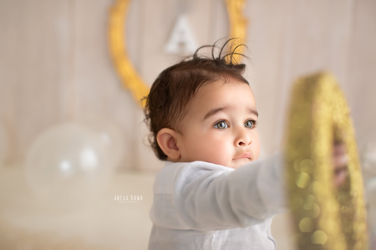 Cheerful boy playing with alphabet balloons beside a golden alphabet frame on the wall, against a serene beige backdrop, capturing the joy of his 12 months first birthday photoshoot in Delhi by Anega Bawa, Gurgaon, Noida
