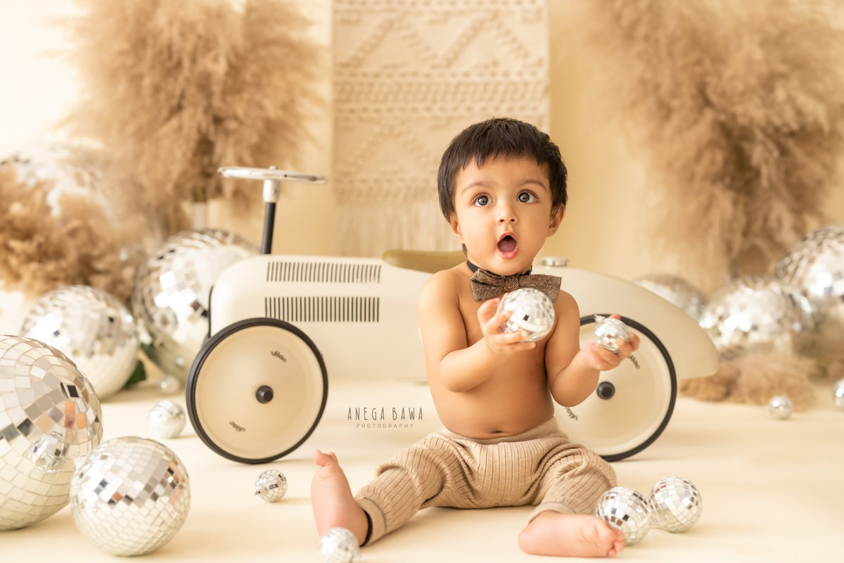 In Gurgaon, Anega Bawa, a skilled sitter photographer, captures the playful spirit of a 12-month-old boy as he joyfully plays with disco balls and a toy scooter against a charming beige backdrop, adorned with a cute bow.
