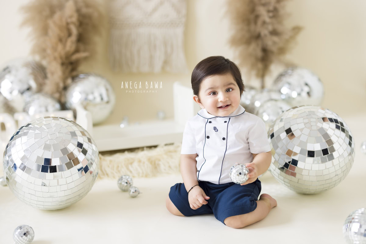 In Gurgaon, Anega Bawa, an expert sitter photographer, captures the playful antics of a 12-month-old boy as he explores and enjoys playing with disco balls amidst brown bushes against a serene beige backdrop.