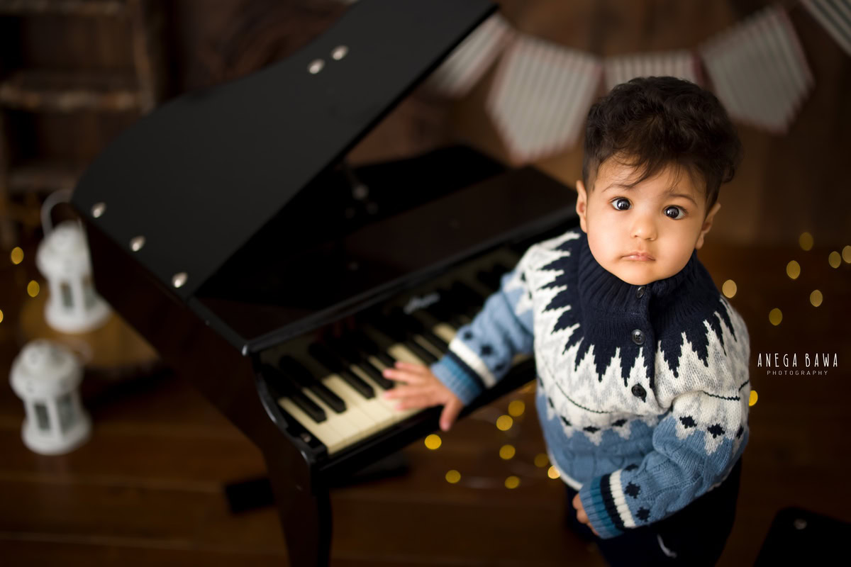 Captured during a memorable sitter photoshoot in Delhi, Gurgaon by Anega Bawa, this adorable 13-month-old boy showcases his musical talents as he plays the piano against a warm brown backdrop. The scene is illuminated by golden lights, adding a magical touch to the moment.