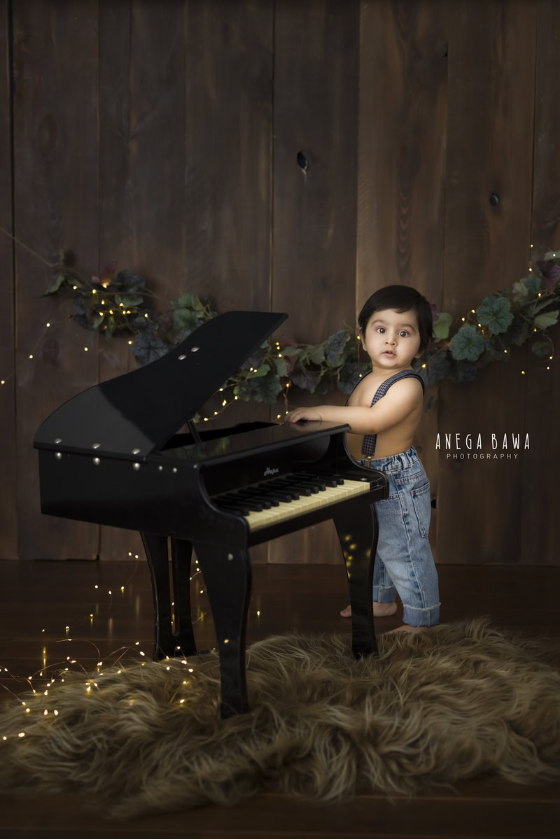 In this enchanting moment captured during a sitter photoshoot in Delhi, Gurgaon by Anega Bawa, a 13-month-old boy is seen playing the piano with joy. Against a rustic brown backdrop and a soft brown rug, fairy lights twinkle, adding a magical ambiance to the scene.