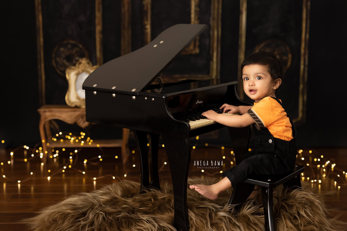 Captured during a sitter photoshoot in Delhi, Gurgaon by Anega Bawa, this adorable 13-month-old boy is immersed in playing the piano. Set against a warm brown backdrop and a cozy brown rug, fairy lights twinkle around him, creating a magical atmosphere.