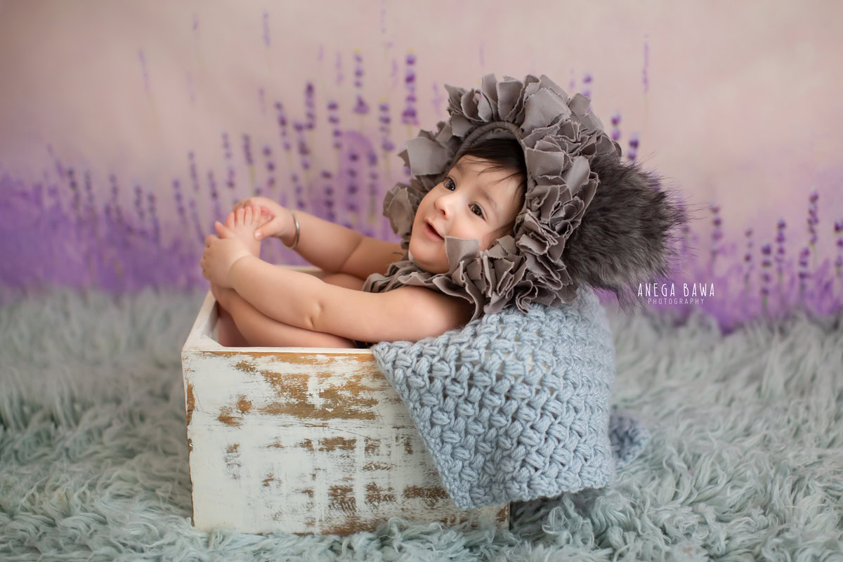 In this enchanting capture from a sitter photoshoot in Delhi, Gurgaon by Anega Bawa, a 13-month-old boy strikes a charming pose from his cozy cot. Adorned with a unique cap, he sits on a soft grey rug against a backdrop blending shades of lavender and pink, creating a serene and captivating scene.