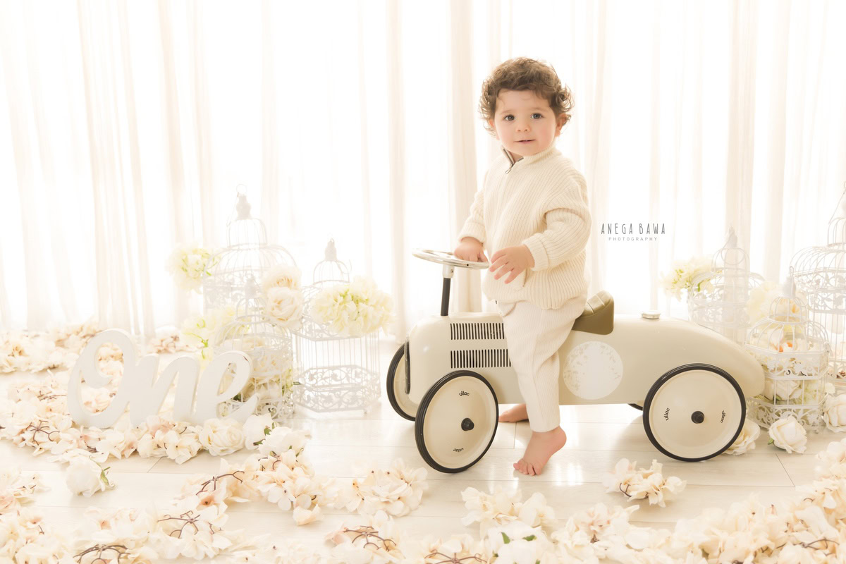 In this delightful scene from a baby pre-birthday photoshoot by Anega Bawa in Delhi, a 13-month-old boy joyfully rides a toy scooter amidst a backdrop of delicate white flowers against a pristine white background. His innocent smile and carefree spirit are beautifully captured, adding a touch of innocence and charm to the moment. It's a heartwarming image that encapsulates the joy and wonder of childhood.