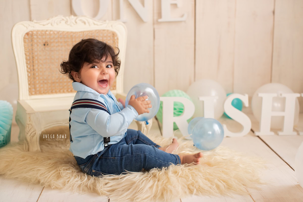 Create lasting memories of your 13-month-old boy with a charming sitter photoshoot by Anega Bawa Photography in Delhi and Gurgaon. Watch him play amidst colorful balloons, seated on a beige rug, with a wooden chair and a name frame adding a personal touch against a serene beige backdrop.