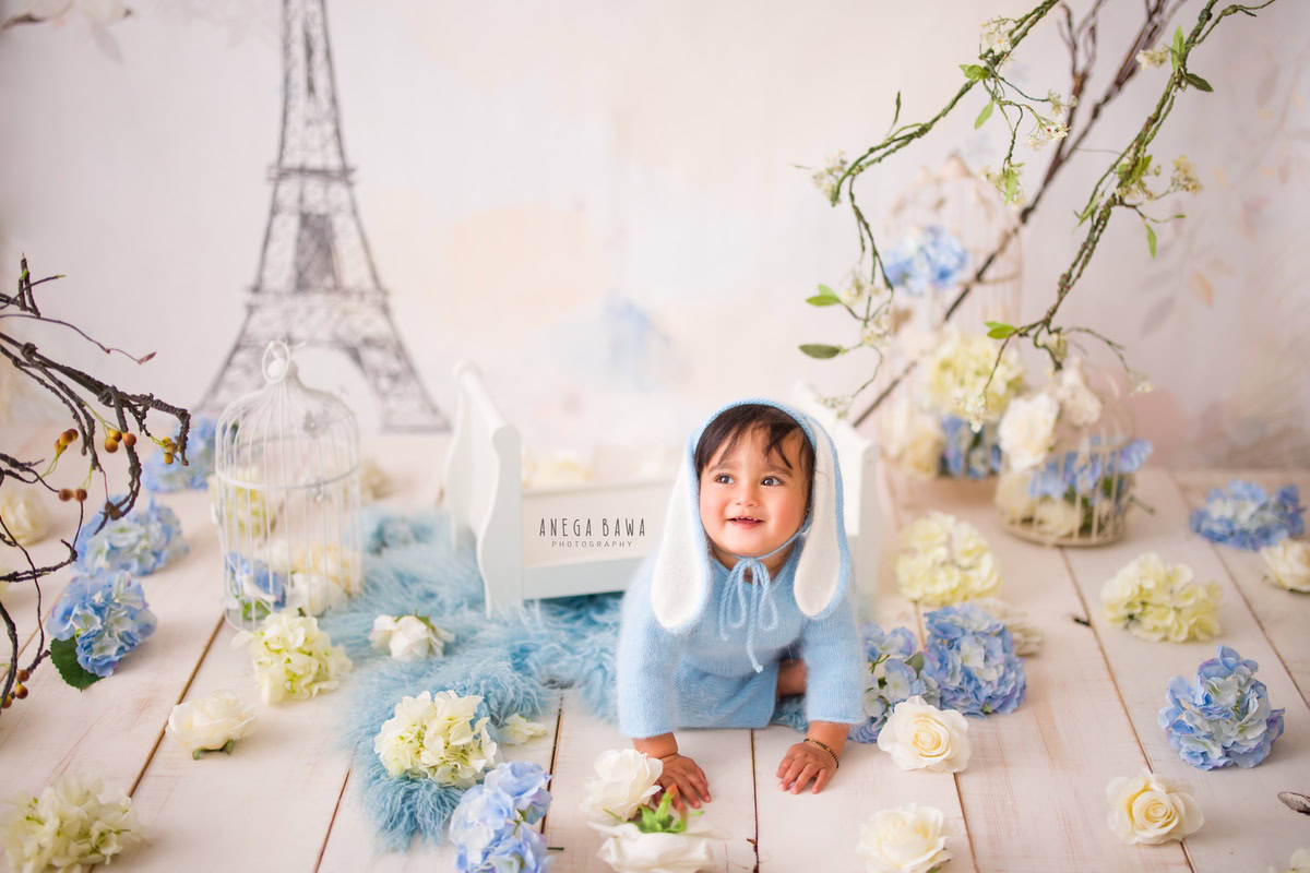 In Gurgaon, Anega Bawa, a talented sitter photographer, captures the charm of a 13-month-old boy seated on a plush blue rug, adorned in a delightful blue and white flowers-themed baby suit and cap. Against the backdrop of an Eiffel Tower wallpaper, the scene exudes a whimsical and enchanting vibe, immortalizing this precious moment in time.