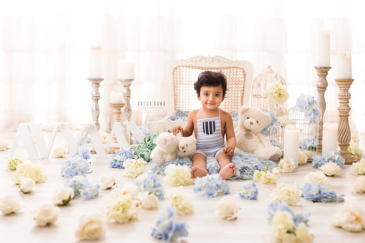 In a delightful scene captured during a sitter photoshoot in Delhi, Gurgaon by Anega Bawa, a 13-month-old boy sits on a cozy blue rug against a pristine white backdrop. Surrounding him are elegant candle frames and adorable blue-and-beige flowers, adding a touch of charm to the serene setting.