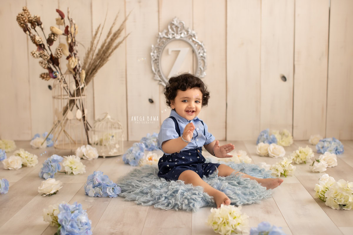 In this captivating moment from a sitter photoshoot in Delhi, Gurgaon by Anega Bawa, a 13-month-old boy sits comfortably on a soft blue rug. Behind him, a silver name frame adorns the wall against a soothing beige backdrop, creating a serene and stylish ambiance.