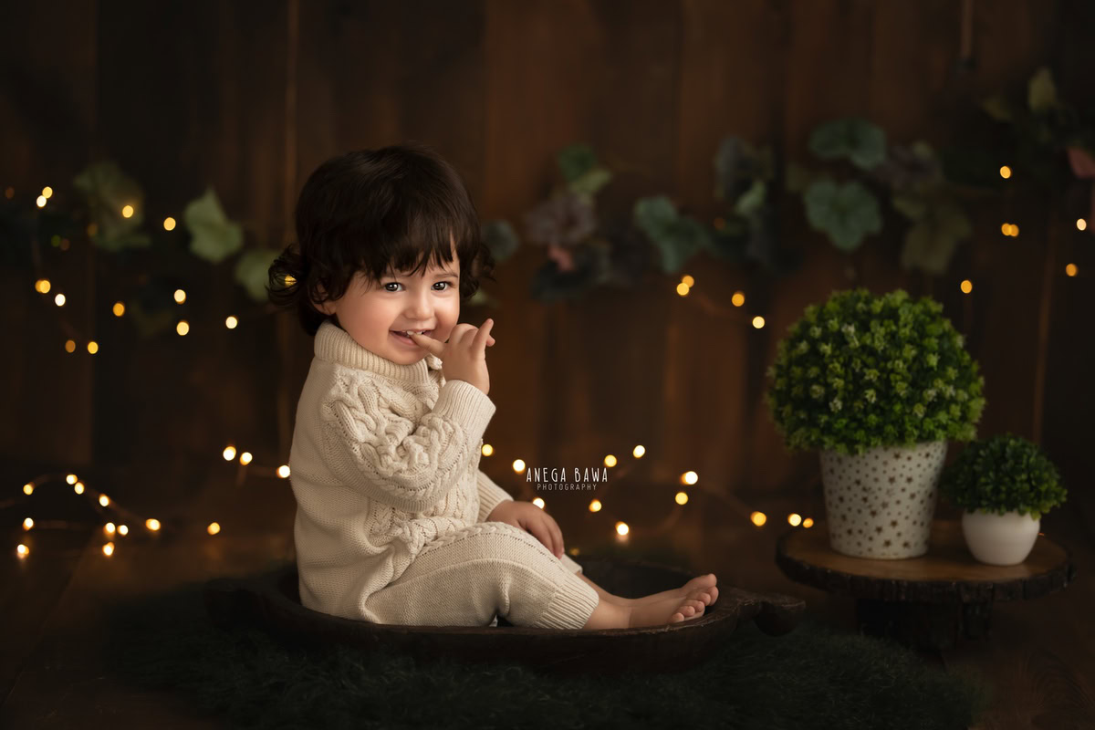 Capture the precious moments of your 13-month-old boy with a delightful sitter photoshoot by Anega Bawa Photography in Delhi and Gurgaon. Set against a warm brown backdrop, adorned with golden lights, watch your little one seated on a cozy brown rug, creating memories to cherish forever.