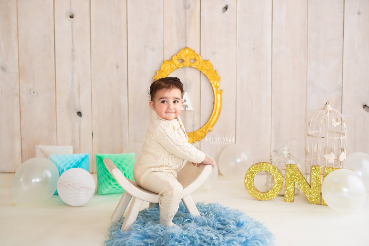 Adorable boy seated on a stylish curvy chair atop a blue rug, surrounded by castles and a golden frame on the wall, against a serene beige backdrop, commemorating his 14 months first birthday photoshoot in Delhi by Anega Bawa, Gurgaon, Noida