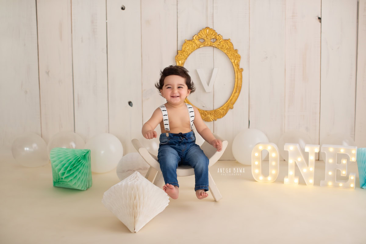 Immerse in the joy of your 14-month-old boy's sitter photoshoot with Anega Bawa Photography in Delhi and Gurgaon. Against a serene beige backdrop adorned with a golden frame on the wall and colorful pompoms, watch your little one seated on a charming curvy chair, capturing moments you'll treasure forever.
