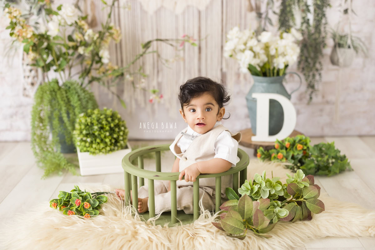 In this enchanting moment captured by Anega Bawa during a baby pre-birthday photoshoot in Delhi, a 14-month-old boy sits comfortably in a green cot against a soothing beige backdrop. Surrounding him are delicate flowers and leaves, adding a natural touch to the scene. In the background, an alphabet frame adds a playful element to the composition. It's a charming image that encapsulates the innocence and wonder of early childhood.