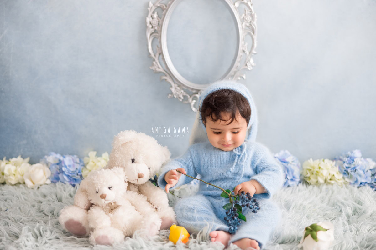 In this delightful sitter photoshoot moment by Anega Bawa in Delhi, Gurgaon, a 14-month-old boy is seated on a cozy grey rug, clad in a charming blue babysuit. Surrounding him, a tender arrangement of blue and white flowers adds a touch of whimsy, while a teddy bear frame on the wall completes the scene against a serene blue backdrop.