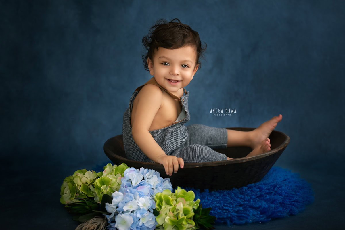 In Gurgaon, Anega Bawa, a skilled sitter photographer, artfully captures the innocence of a 14-month-old boy seated in a tub, surrounded by a serene blue backdrop and adorned with vibrant blue flowers. This captivating scene, set against a soothing blue rug, perfectly encapsulates the joy and wonder of childhood, creating a timeless keepsake for families to cherish.