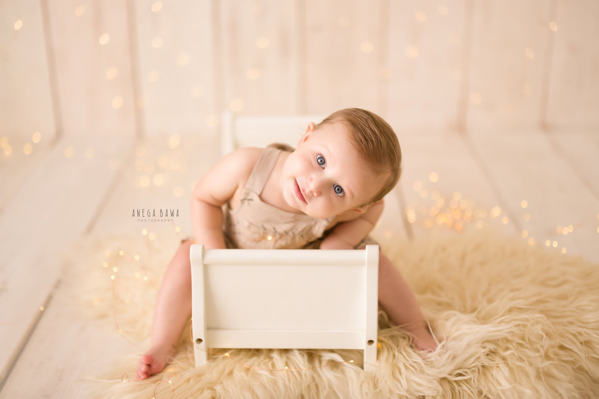 Captured in the enchanting sitter photoshoot by Anega Bawa in Delhi, Gurgaon, this 14-month-old boy sits serenely in a white cot adorned with a beige rug. The scene is illuminated by the soft glow of fairy lights, creating a magical ambiance for this adorable moment.