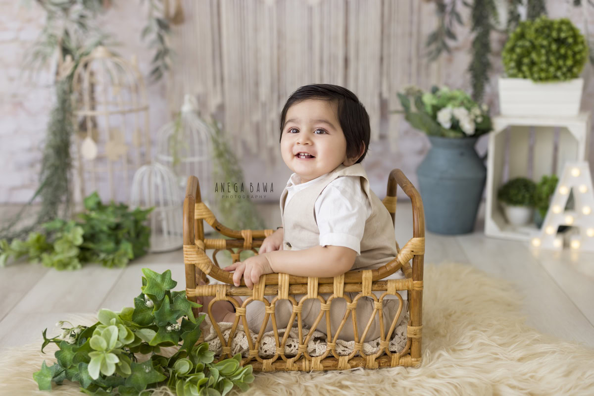 Captured by Anega Bawa during a delightful baby pre-birthday photoshoot in Delhi, this image features a 14-month-old boy seated snugly in a wooden cot. The beige rug beneath him offers a soft and comforting touch, while nearby, an alphabet frame adds a playful element. Blue pots and a bunch of leaves on the floor create a natural and vibrant atmosphere, enhancing the charm of the scene. It's a captivating moment that reflects the innocence and joy of early childhood.