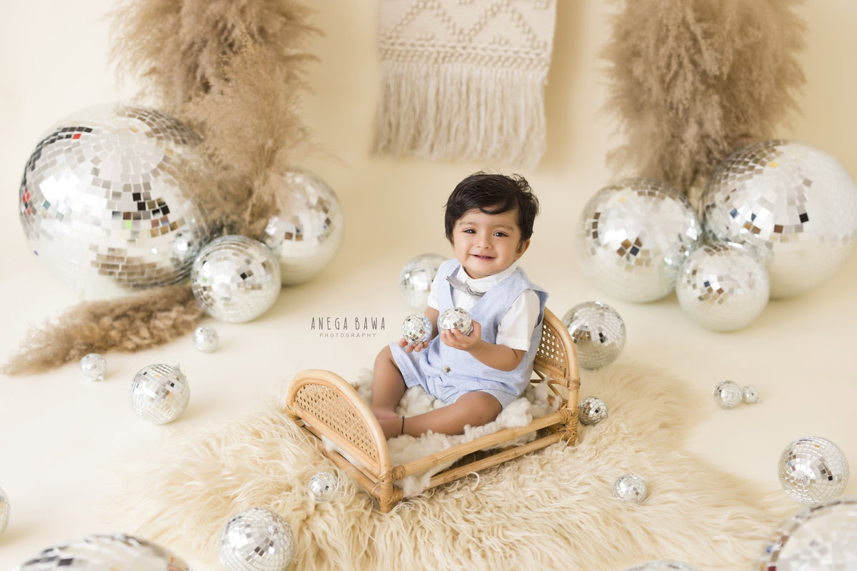 Adorable boy seated in a wooden cot on a cozy beige rug amidst lush bushes and sparkling disco balls against a serene beige backdrop, commemorating his 14 months first birthday photoshoot in Delhi by Anega Bawa, Gurgaon, Noida