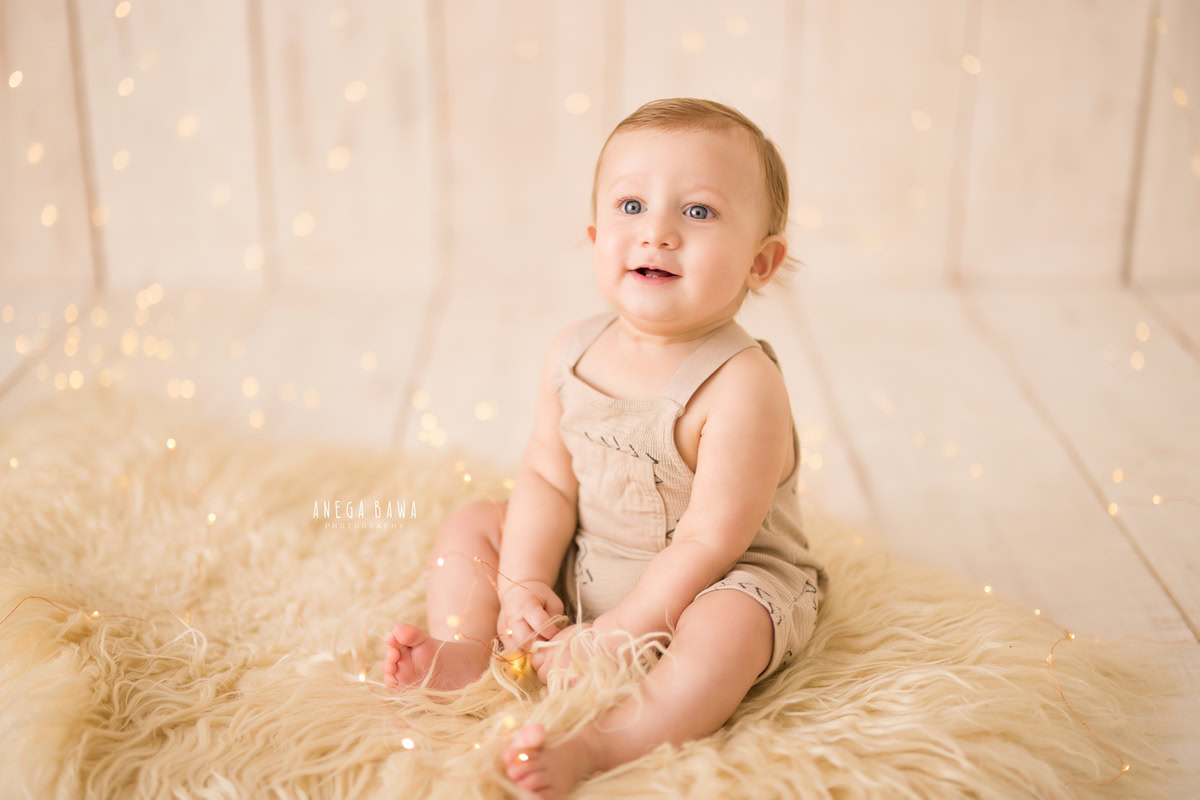 In Gurgaon, Anega Bawa, a talented sitter photographer, beautifully captures the charm of a 14-month-old boy seated on a cozy beige rug against a serene beige backdrop. The warm glow of golden lights adds a magical touch to the scene, creating a captivating portrait that exudes comfort and tranquility.