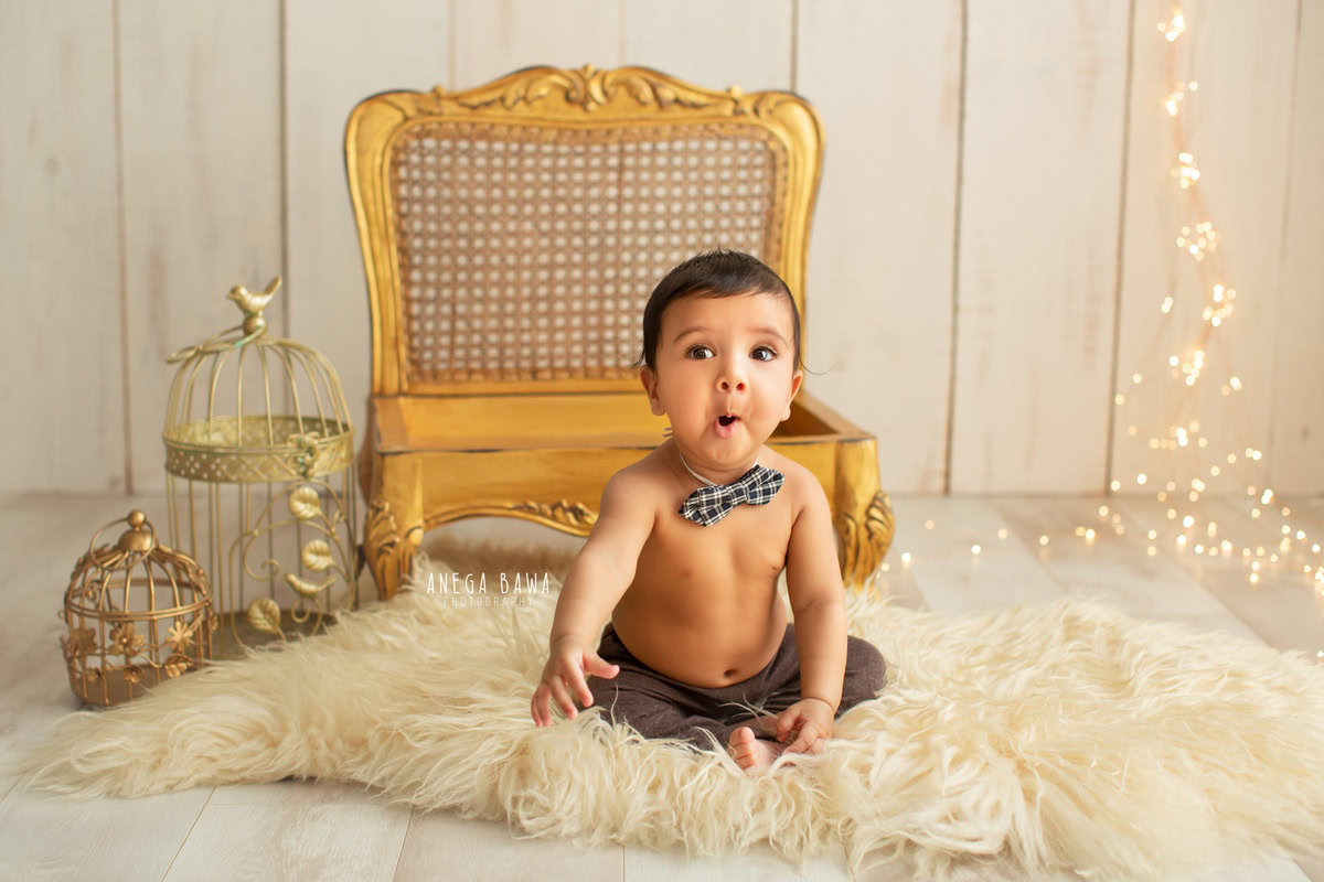 In Gurgaon, Anega Bawa, a skilled sitter photographer, captures the innocence and charm of a 14-month-old boy seated on a luxurious beige rug, adorned with a golden chair and castles. Against a soft beige backdrop, the scene is elevated with a delightful bow, creating a captivating portrait filled with warmth and elegance.