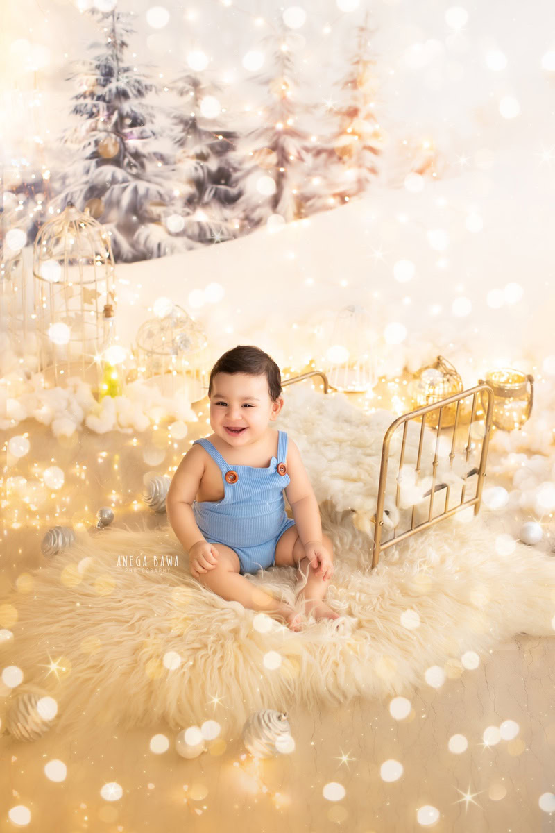 Captivating boy seated on a cozy beige rug beside a golden cot, surrounded by twinkling fairy lights against a pristine white backdrop, commemorating his 14 months first birthday photoshoot in Delhi by Anega Bawa, Gurgaon, Noida