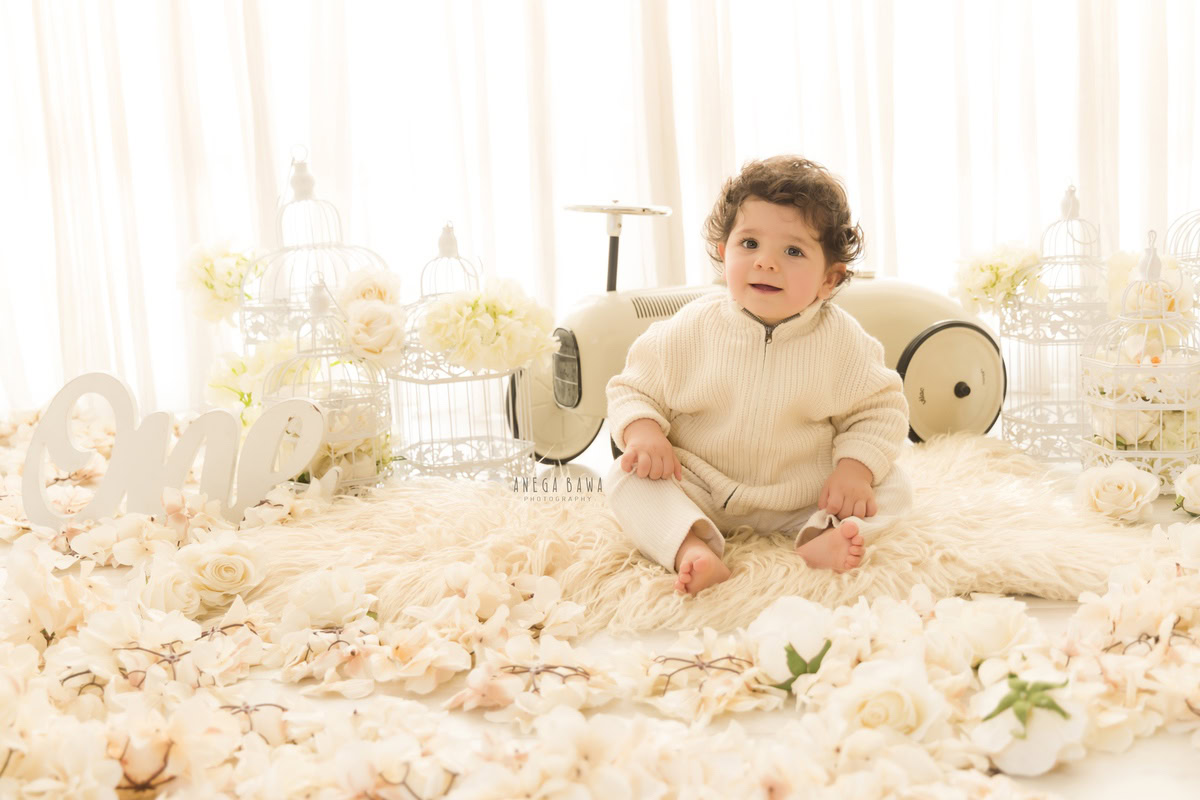 Adorable boy seated on a cozy beige rug surrounded by white castles and a toy scooter against a pristine white backdrop, commemorating his 14 months first birthday photoshoot in Delhi by Anega Bawa, Gurgaon, Noida