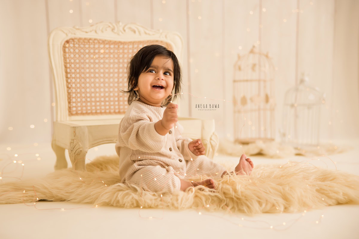 Delight in your 14-month-old boy's sitter photoshoot by Anega Bawa Photography in Delhi and Gurgaon. Against a warm beige backdrop adorned with white castles, witness your little one seated on a wooden chair on a soft beige rug, capturing precious moments you'll cherish forever.