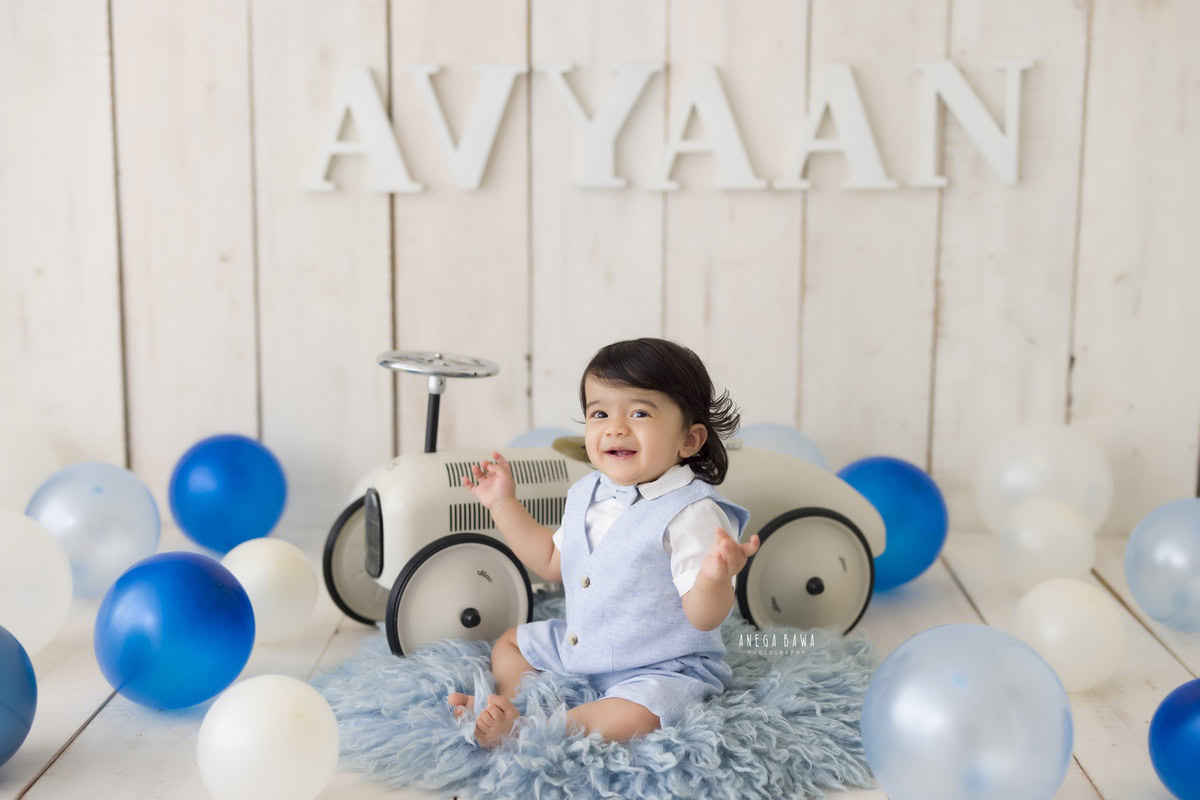 Delight in the memories of your 15-month-old boy with Anega Bawa Photography's sitter photography in Delhi and Gurgaon. Picture him seated on a blue rug, exploring his world on a toy scooter amidst colorful balloons. The name frame on the wall adds a personal touch against the soothing beige backdrop, creating timeless moments to cherish.