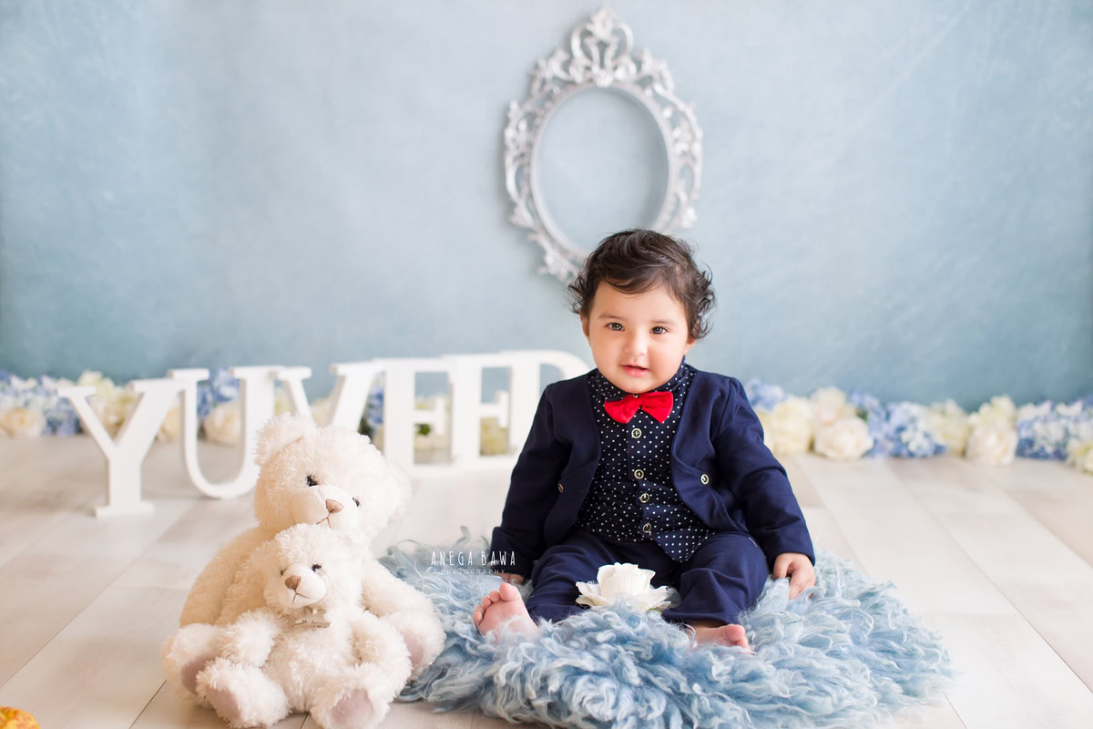 Celebrate your 15-month-old boy's sitter milestone with Anega Bawa Photography in Delhi and Gurgaon. Adorned in a charming blue coat and pants with a red bow, he sits on a cozy blue rug amidst teddy bears, against a serene blue backdrop complemented by a silver frame on the wall, capturing precious memories to cherish for years to come.
