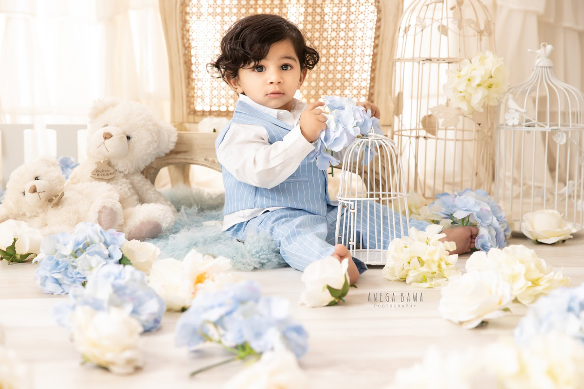 In Gurgaon, Anega Bawa, an adept sitter photographer, skillfully captures the serenity of a 15-month-old boy seated on a plush blue rug amidst a scene of white castles, soft toys, and scattered flowers on the floor. Against a soothing beige backdrop, the composition exudes a tranquil charm, evoking a sense of wonder and innocence in the young subject.