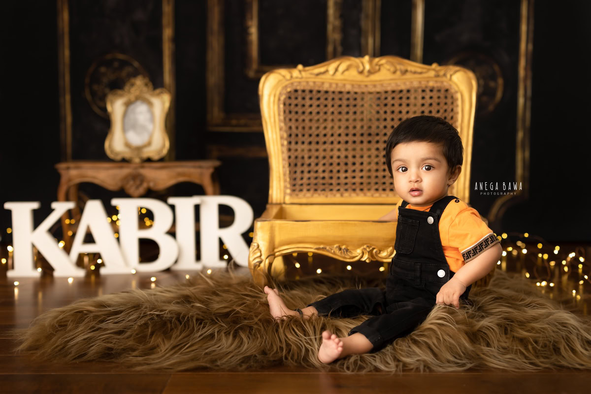 In this captivating sitter photography session by Anega Bawa in Delhi, Gurgaon, the 15-month-old boy sits gracefully on a brown rug. His regal presence is accentuated by a golden chair, while a name frame on the floor adds a personal touch against the warm brown backdrop.