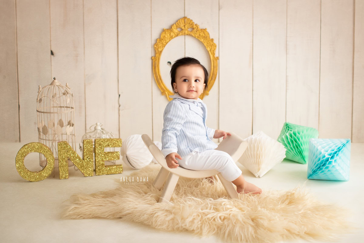 Captivating boy seated on a stylish curvy chair against a serene beige backdrop, surrounded by charming white castles and colorful pompoms, celebrating his 15 months first birthday photoshoot in Delhi by Anega Bawa, Gurgaon, Noida