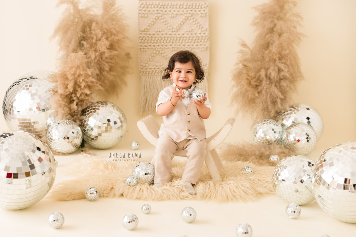 In this charming sitter photography session by Anega Bawa in Delhi, Gurgaon, the 15-month-old boy sits on a curvy chair placed on a beige rug. The scene is illuminated with disco balls, and brown bushes add a natural touch against the serene beige backdrop.
