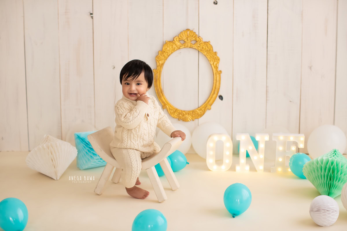 In Gurgaon, Anega Bawa, a talented sitter photographer, frames a captivating moment of a 15-month-old boy seated on a curvy chair. The scene is adorned with a golden frame on the wall and vibrant balloons against a warm beige backdrop. With expert precision, the photographer captures the essence of childhood innocence and joy in this delightful composition.