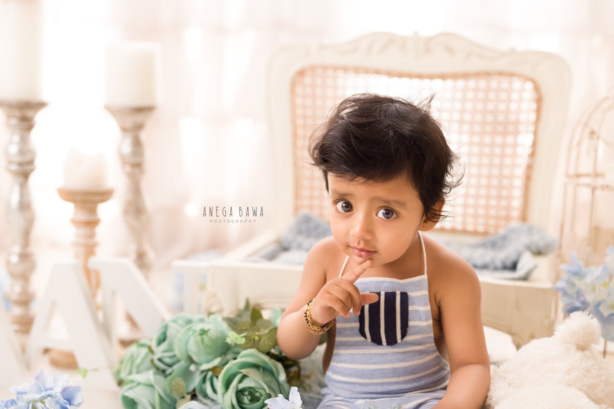 In this delightful sitter photography session by Anega Bawa in Delhi, Gurgaon, a 15-month-old boy is captured seated on the floor amidst green flowers. A wooden chair and a candle stand add to the charming scene against a beige backdrop.