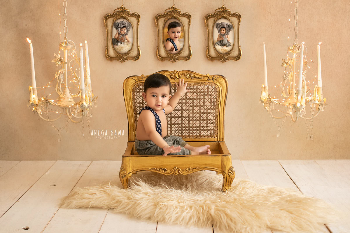 In this enchanting sitter photography session by Anega Bawa in Delhi, Gurgaon, a 15-month-old boy is captured seated on a golden chair beside a candle stand and adorned with golden lights. The scene is further enhanced by photo frames on the wall against a beige backdrop.