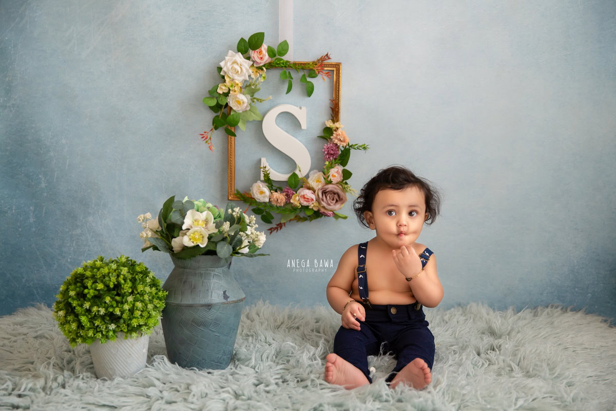 In this delightful sitter photography session by Anega Bawa in Delhi, Gurgaon, a 15-month-old boy is captured seated on a grey rug, surrounded by a charming arrangement. Behind him, a floral frame adorns the wall, complementing the blue backdrop. Additionally, flower pots add a touch of natural beauty to the scene.