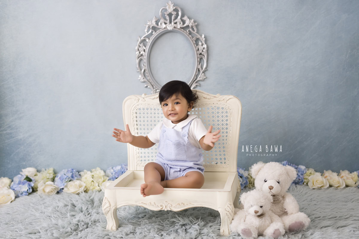 Adorable boy seated on a white wooden chair against a sophisticated grey backdrop, accompanied by a soft toy on a cozy grey rug, celebrating his 15 months first birthday photoshoot in Delhi by Anega Bawa, Gurgaon, Noida